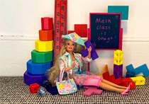 A Barbie doll sitting down on the floor, with a range of mathematical blocks and tools behind her. There is also a blackboard with the words 'Math class is tough!' written on it.