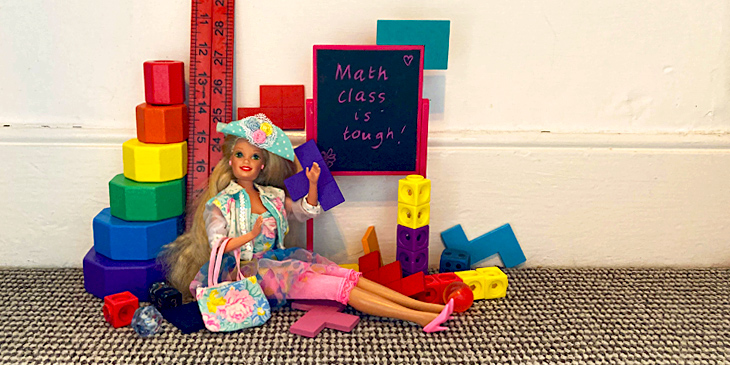 A Barbie doll sitting down on the floor, with a range of mathematical blocks and tools behind her. There is also a blackboard with the words 'Math class is tough!' written on it.