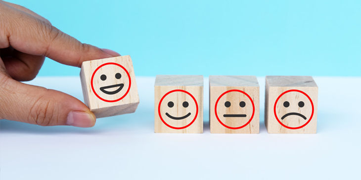 4 wooden blocks with drawn faces on each, ranging from a smile to a frown. A hand is picking up the smiling face