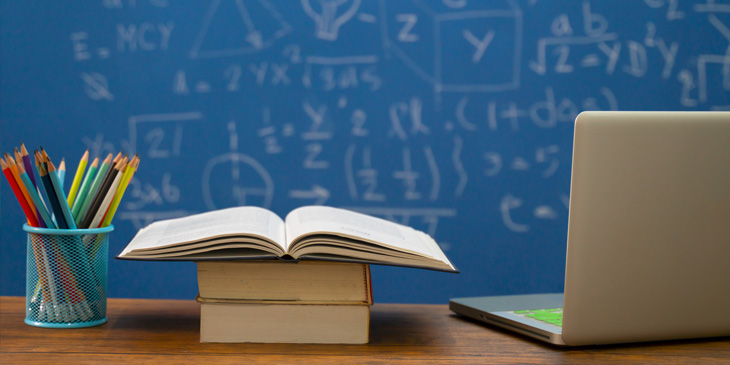 A desk in-front of a blackboard with sums. On the desk is a pencil pot, books, and a laptop