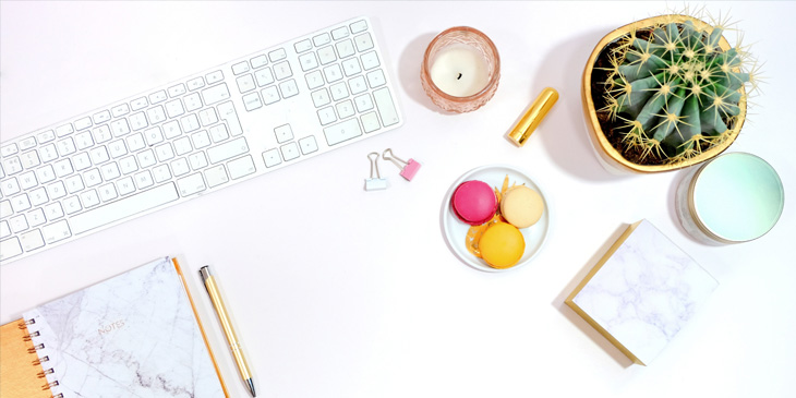 A computer desk with a keyboard, cactus, candle and notepad scattered over it