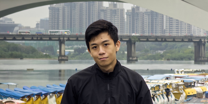 John standing on the edge of a river, with a bridge in the background and a cityscape further behind in the distance