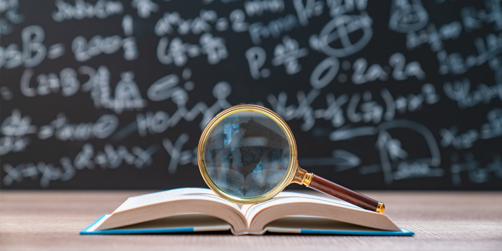 An open book resting on a table with a magnifying glass on top of it. In the background is a blackboard with mathematics equations on.