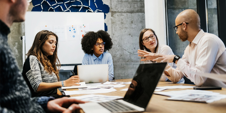 A group of three young women and two men of different ethnicities are in a business meeting in a modern day office